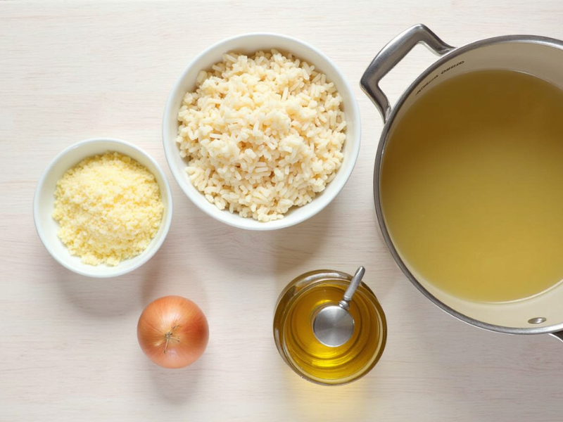 Zutaten für Risotto Rezept: Arborio-Reis, Parmesan, Zwiebel, Brühe und Olivenöl auf einer Arbeitsfläche arrangiert